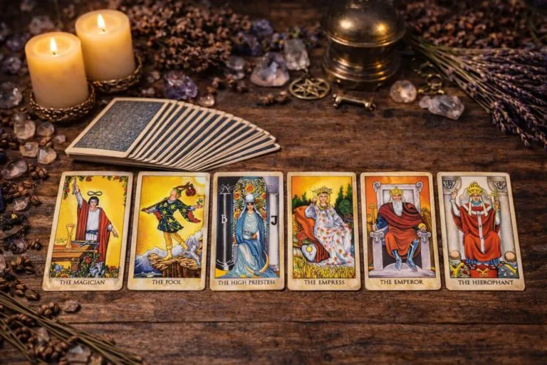 Seis cartas de los Arcanos Mayores del tarot Rider-Waite —El Mago, El Loco, La Sacerdotisa, La Emperatriz, El Emperador y El Hierofante— sobre una mesa rústica con velas, cristales y el mazo de tarot al fondo.